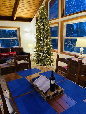 A cozy room with a wooden ceiling, a Christmas tree, a dining table with a bottle of wine, and a view of snow-covered trees through the windows.