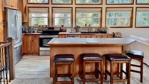 Une cuisine spacieuse dotée de meubles en bois, d’un grand îlot central avec des tabourets et de nombreuses fenêtres offrant une vue sur l’extérieur.