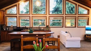 Une cuisine spacieuse avec un îlot central, des chaises en bois, une table à manger et un canapé blanc. De grandes fenêtres offrent une vue sur la forêt environnante.