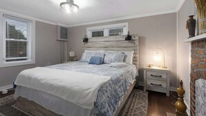 A bedroom with a bed, bedside tables, a nightstand, a lamp, a window with blinds, and a wall-mounted shelf.
