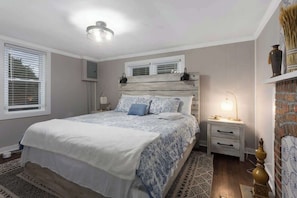 A bedroom with a bed, bedside tables, a nightstand, a lamp, a window with blinds, and a wall-mounted shelf.