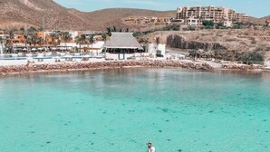 A clear turquoise sea with a person paddleboarding, a resort building, and a hilly landscape in the background. - Quinta with ocean and golf course panoramic views (La Paz)