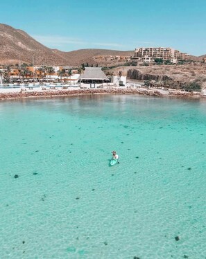 A clear turquoise sea with a person paddleboarding, a resort building, and a hilly landscape in the background.