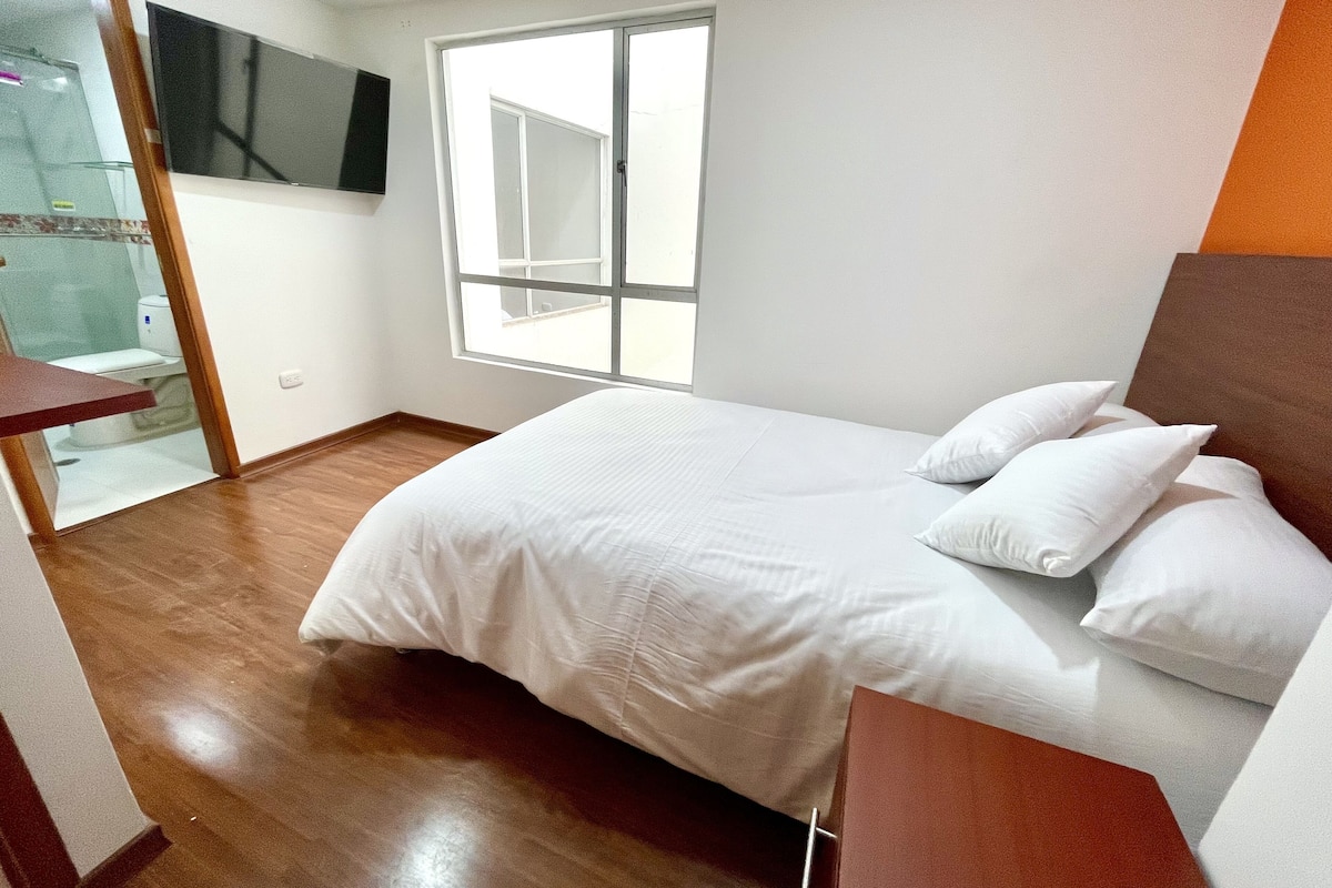 Een slaapkamer met een bed, witte kussens, een houten vloer, een aan de muur bevestigde televisie en een raam.