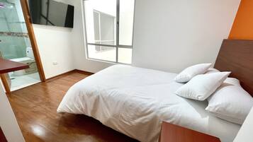 A bedroom with a bed, white pillows, a wooden floor, a TV mounted on the wall, and a window.