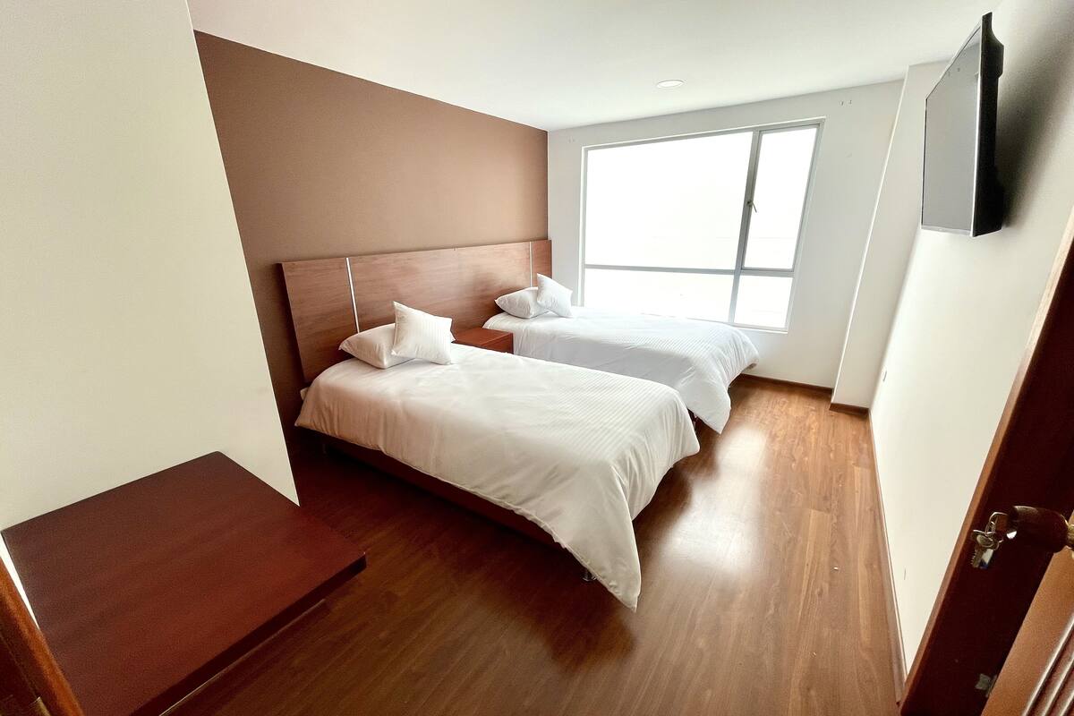Een slaapkamer met twee bedden, een houten vloer, een groot raam en een aan de muur bevestigde televisie.