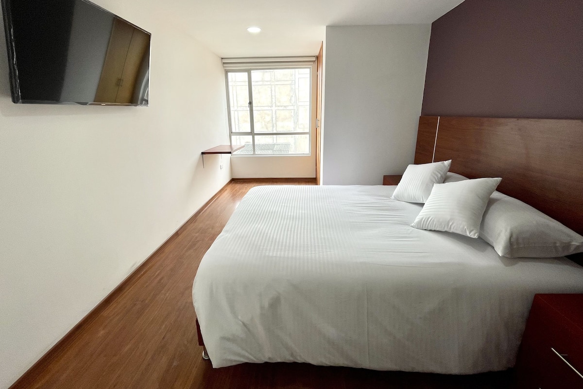 Een slaapkamer met een groot bed, een wandgemonteerde televisie, een raam en een houten vloer.
