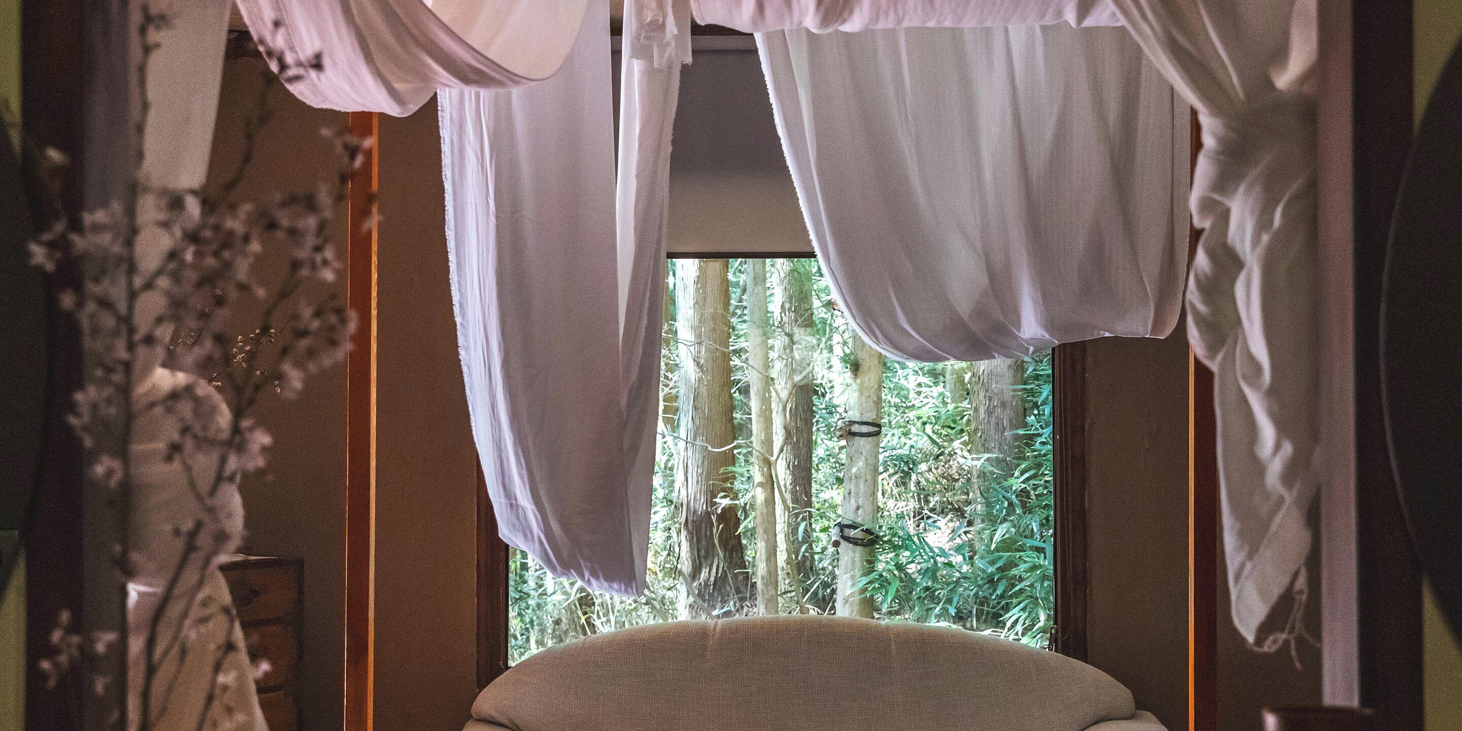 Ein Zimmer mit Himmelsbett, einem mit Vorhängen versehenen Fenster und Blick ins Grüne.