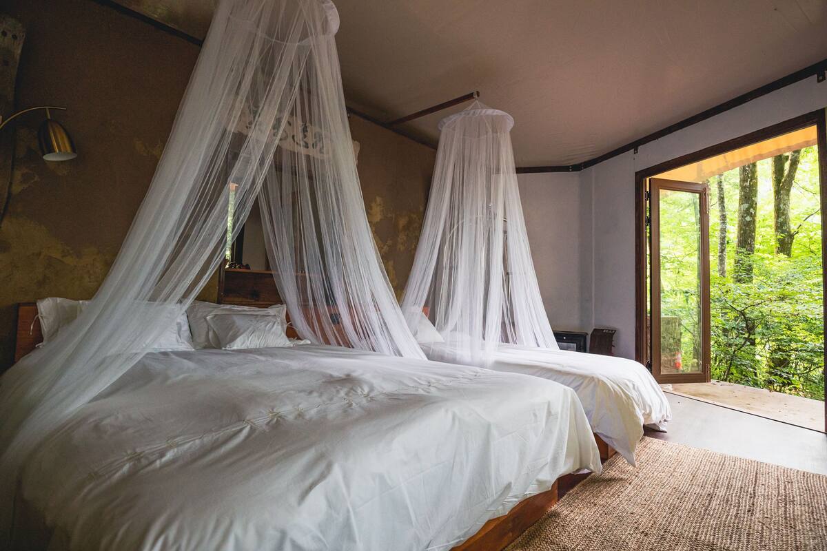Quarto com duas camas, cada uma com dossel. Há uma janela grande que oferece vista para o verde lá fora.
