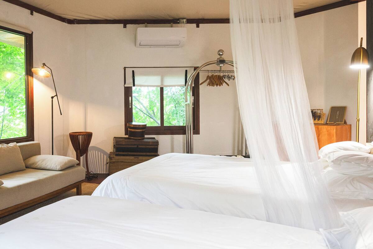 Quarto com duas camas, um sofá, uma janela com vista para árvores, uma luminária e um ar condicionado.