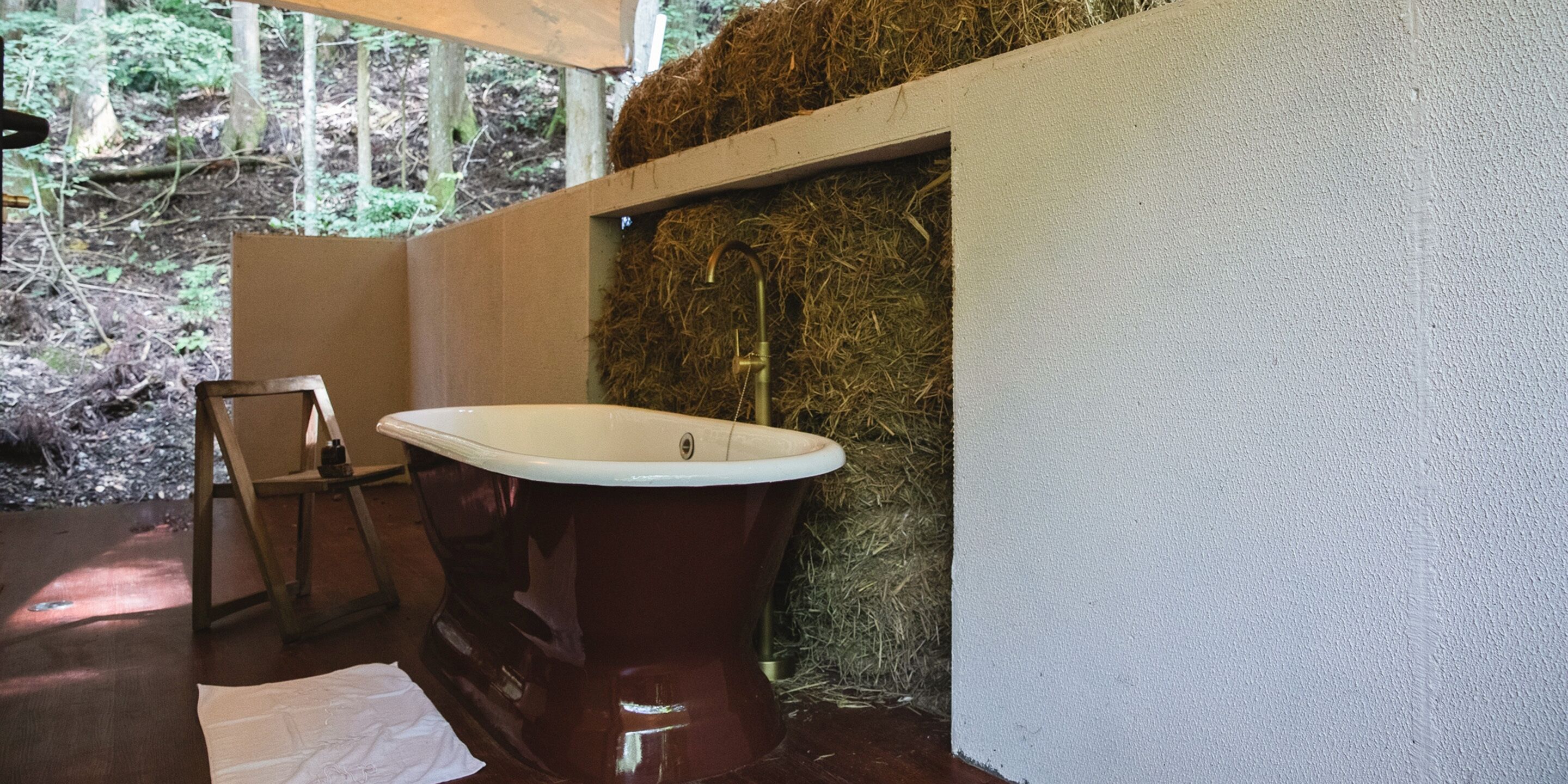 A bathroom with a large, vintage-style bathtub, a wooden chair, and a wall covered in hay.
