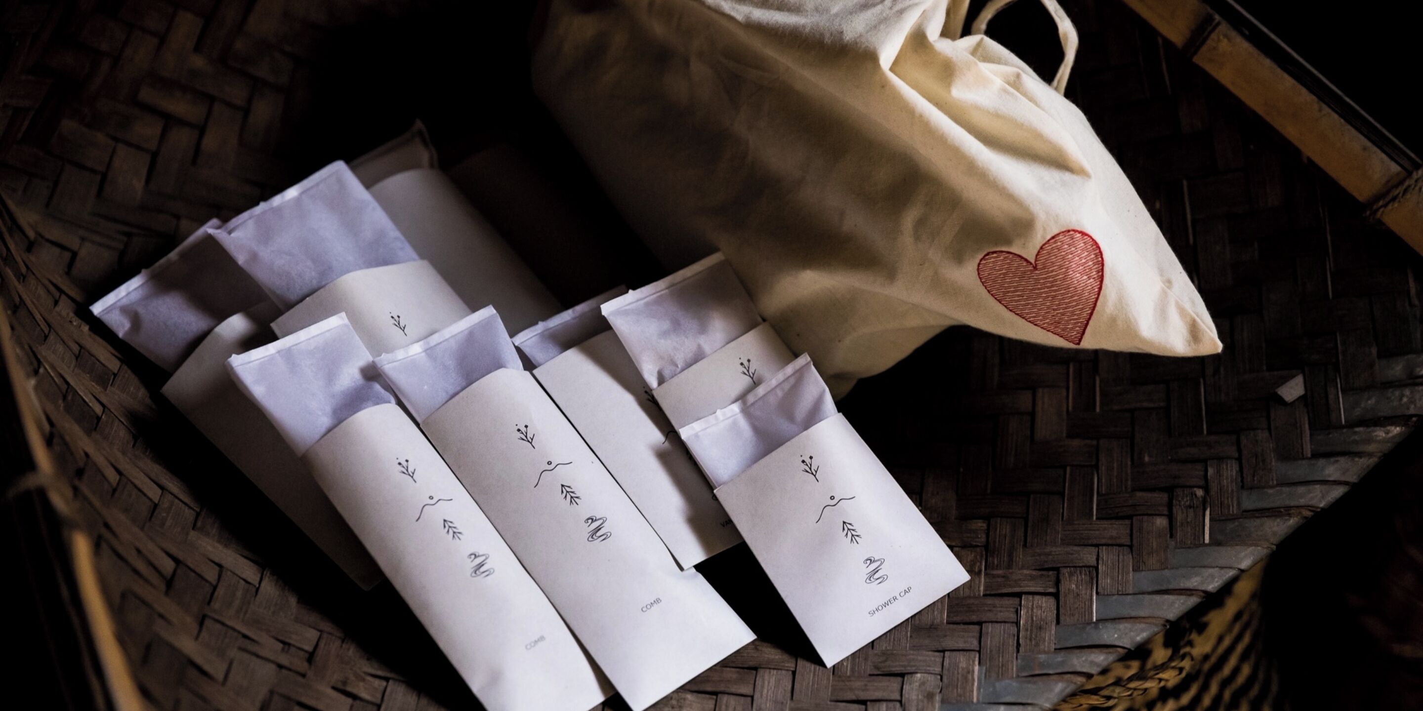 A woven basket containing several white paper bags with embroidered designs and a white tote bag with a heart design.