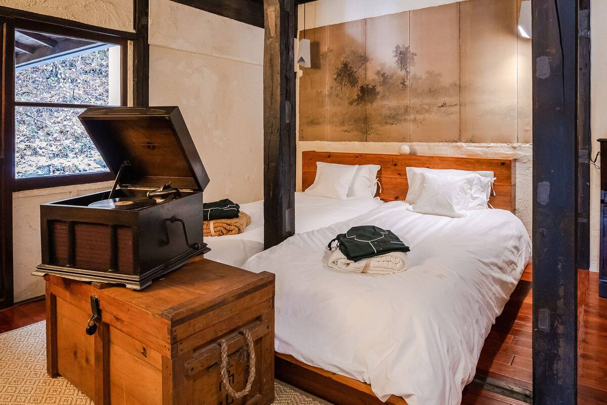 Quarto com um baú de madeira, cama com roupa de cama branca e vista para uma paisagem montanhosa pela janela.