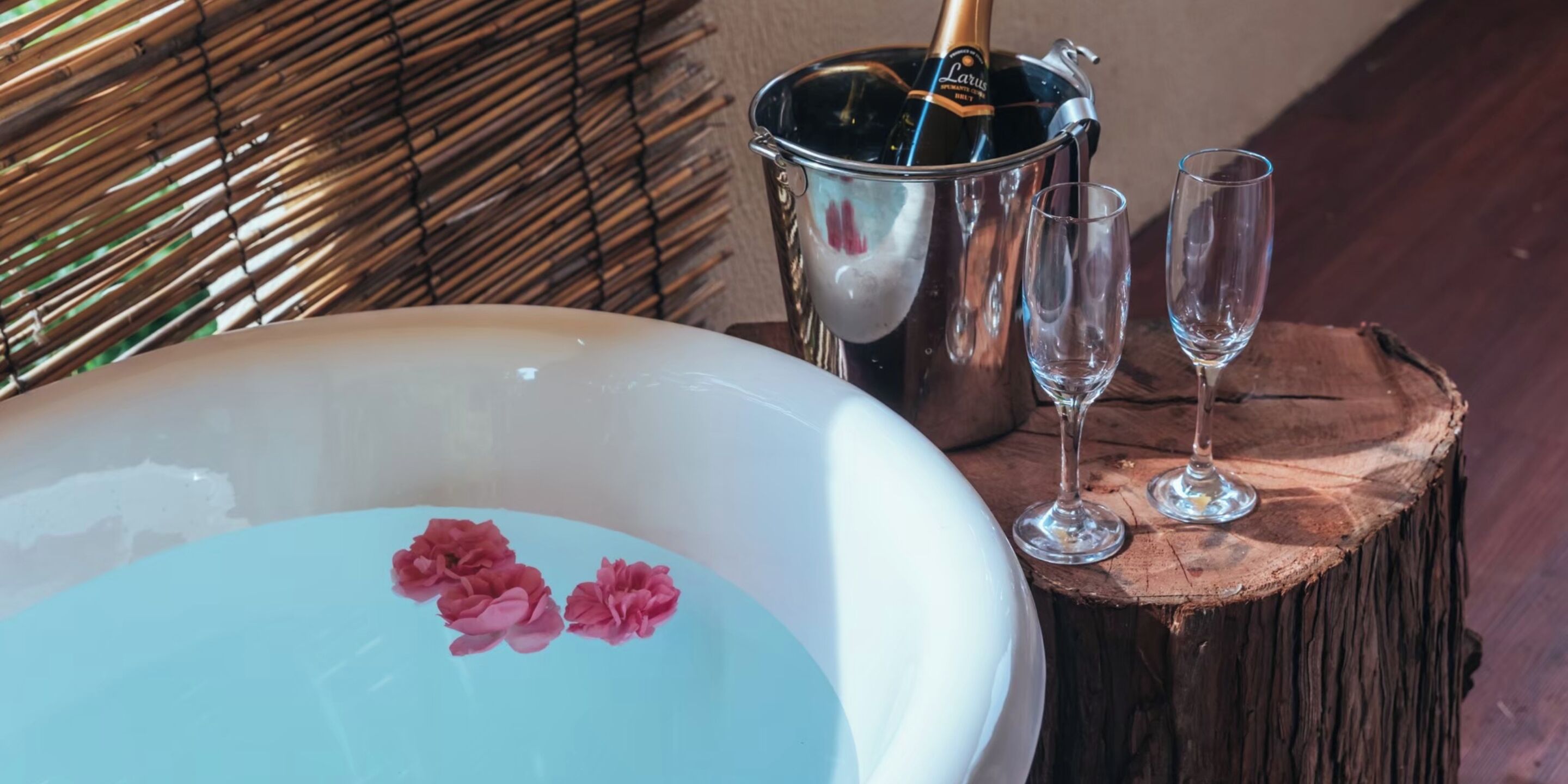 A bathtub with pink flowers, a champagne bottle, and two glasses on a wooden stump.