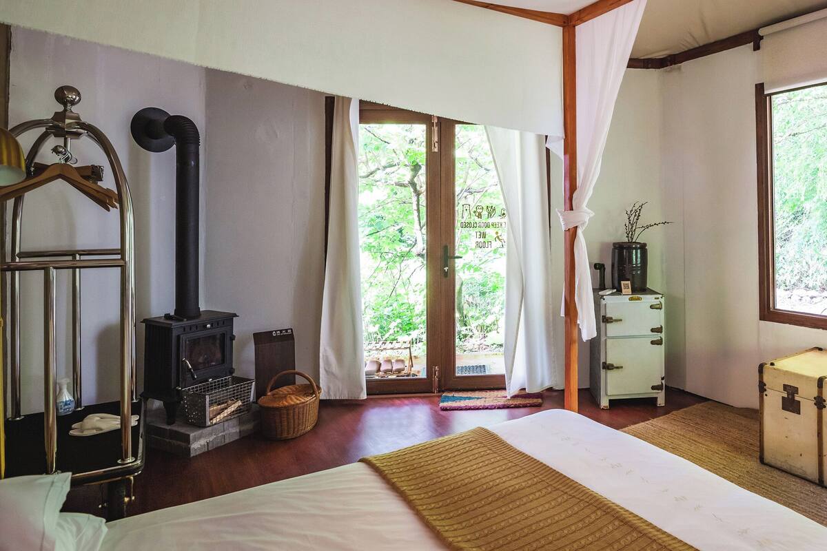 Quarto com cama, piso de madeira, mala, armário pequeno e janela com vista para o verde.