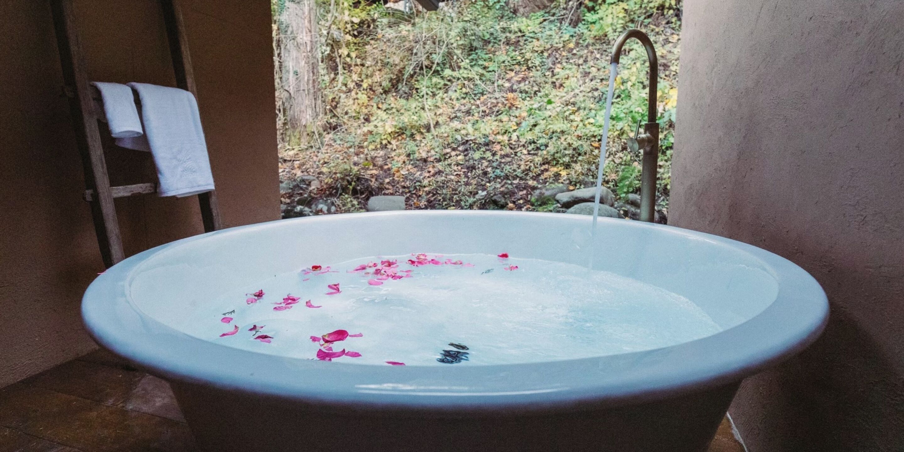 Uma banheira com pétalas de rosas, um porta-toalhas e vista para um jardim.