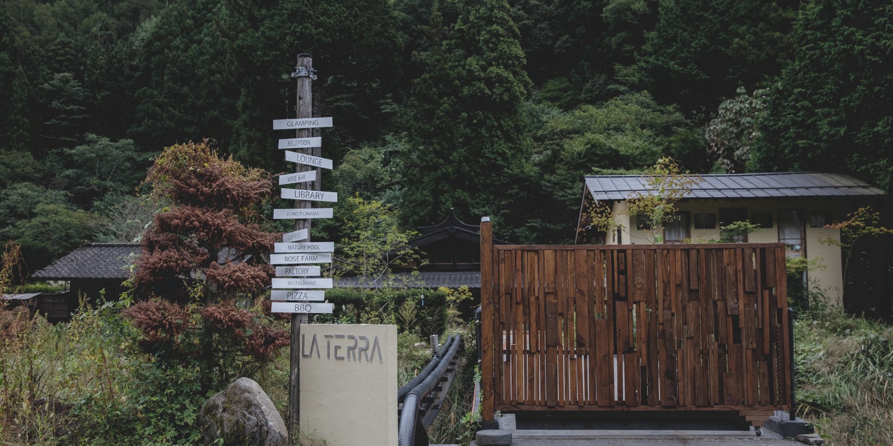 A signpost with multiple directional signs pointing to various amenities such as a glamping site, reception, lounge, wine bar, library, forest cinema, nature pool, rose farm, factory, restaurant, pizza, and BBQ.