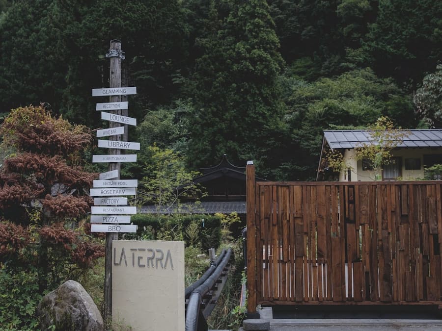 A signpost with multiple directional signs pointing to various amenities such as a glamping site, reception, lounge, wine bar, library, forest cinema, nature pool, rose farm, factory, restaurant, pizza, and BBQ.