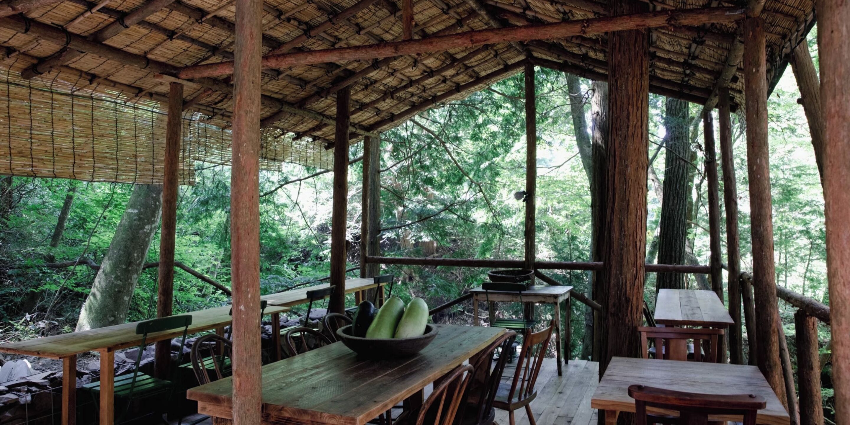 Una estructura de madera rústica con mesas y sillas, rodeada de un bosque frondoso.