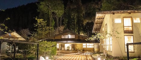 Ein traditionelles japanisches Gebäude mit Holzwänden und einem Ziegeldach, umgeben von Bäumen und einem steinbelagten Weg zum Eingang.