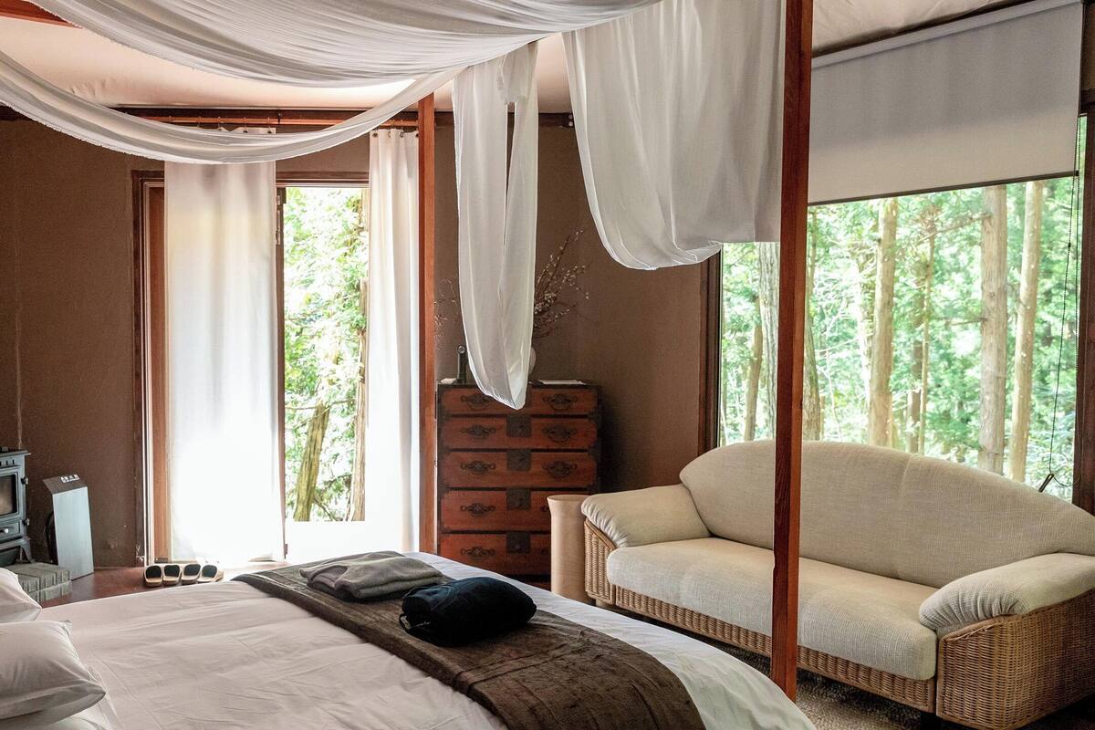Quarto com cama, uma cadeira, cômoda e janela com vista para árvores.
