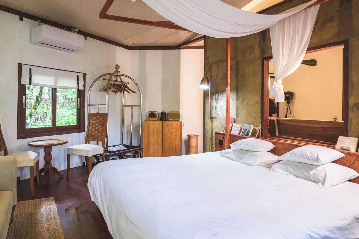 Quarto com uma cama grande, cabeceira de madeira, uma mesinha, uma cadeira e uma janela com vista para o verde.