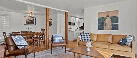 A modern living room with a brown leather sofa, wooden coffee table, and a dining area with a wooden table and chairs.