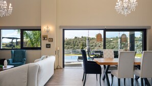 A modern living room with a sofa, a dining table with chairs, and a chandelier. There are large windows offering a view of greenery and a distant hill. - Heated pool, kids pool, gorge and sea views! (Rethymno)