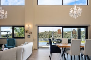 Un salon moderne avec un canapé, une table à manger et des chaises, ainsi qu’un lustre. De grandes fenêtres offrent une vue sur la verdure et une colline lointaine.