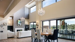 A spacious living area with a fireplace, a dining table with chairs, and large windows offering a view of the outdoors. - Heated pool, kids pool, gorge and sea views! (Rethymno)
