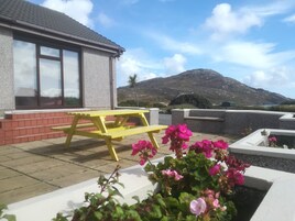 Un patio con una mesa de picnic amarilla y flores rosadas en macetas, un edificio con ventanas grandes y un telón de fondo montañoso.