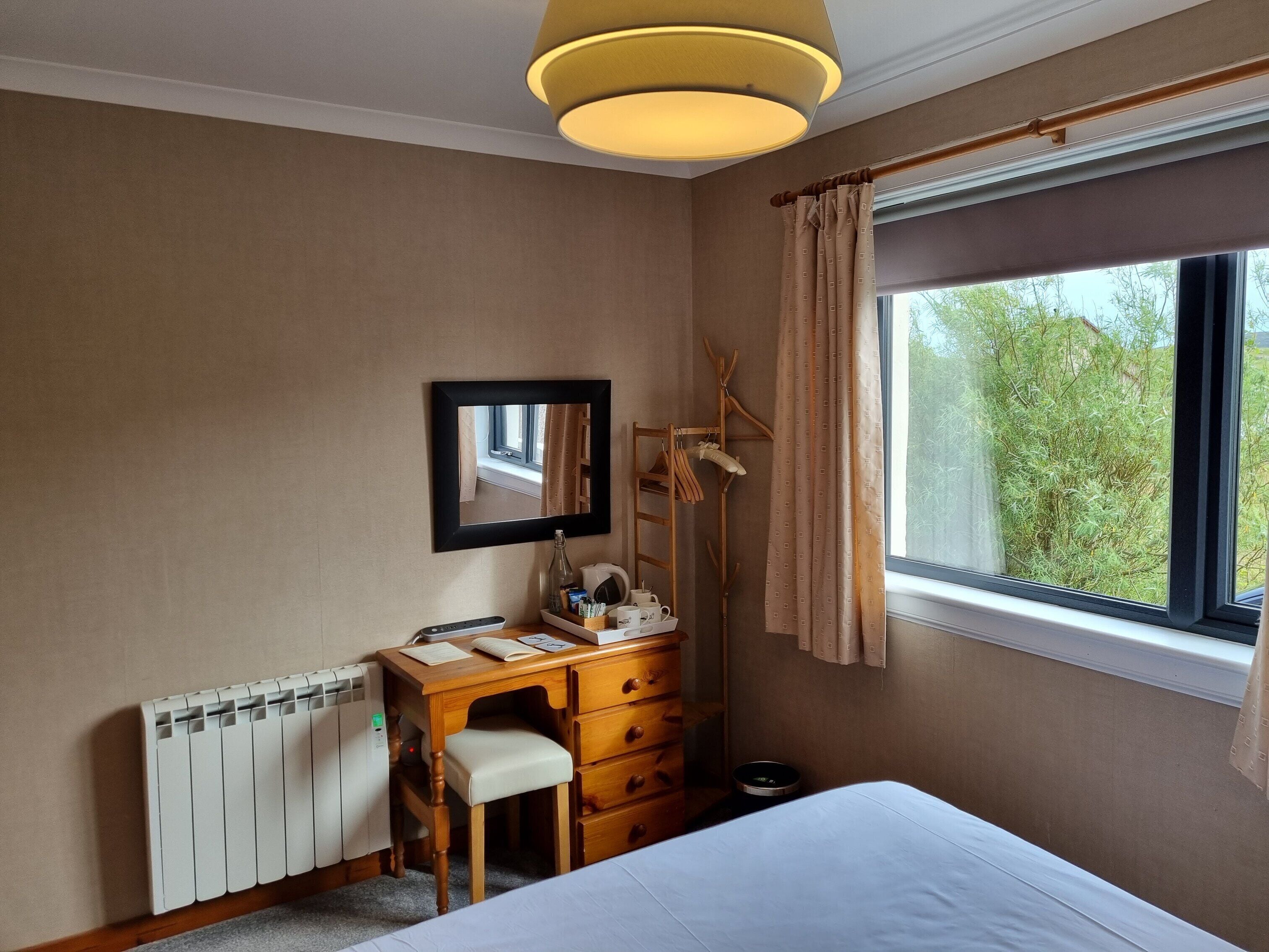 Une chambre d’hôtel avec un bureau en bois, une chaise, un lit, un radiateur, une fenêtre avec des rideaux et un porte-manteau mural.