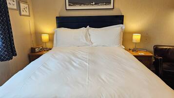Un lit bien fait, avec du linge de lit blanc, une tête de lit sombre et deux lampes de chevet. Un tableau encadré est accroché au-dessus du lit, et un manteau est suspendu au mur, à gauche.