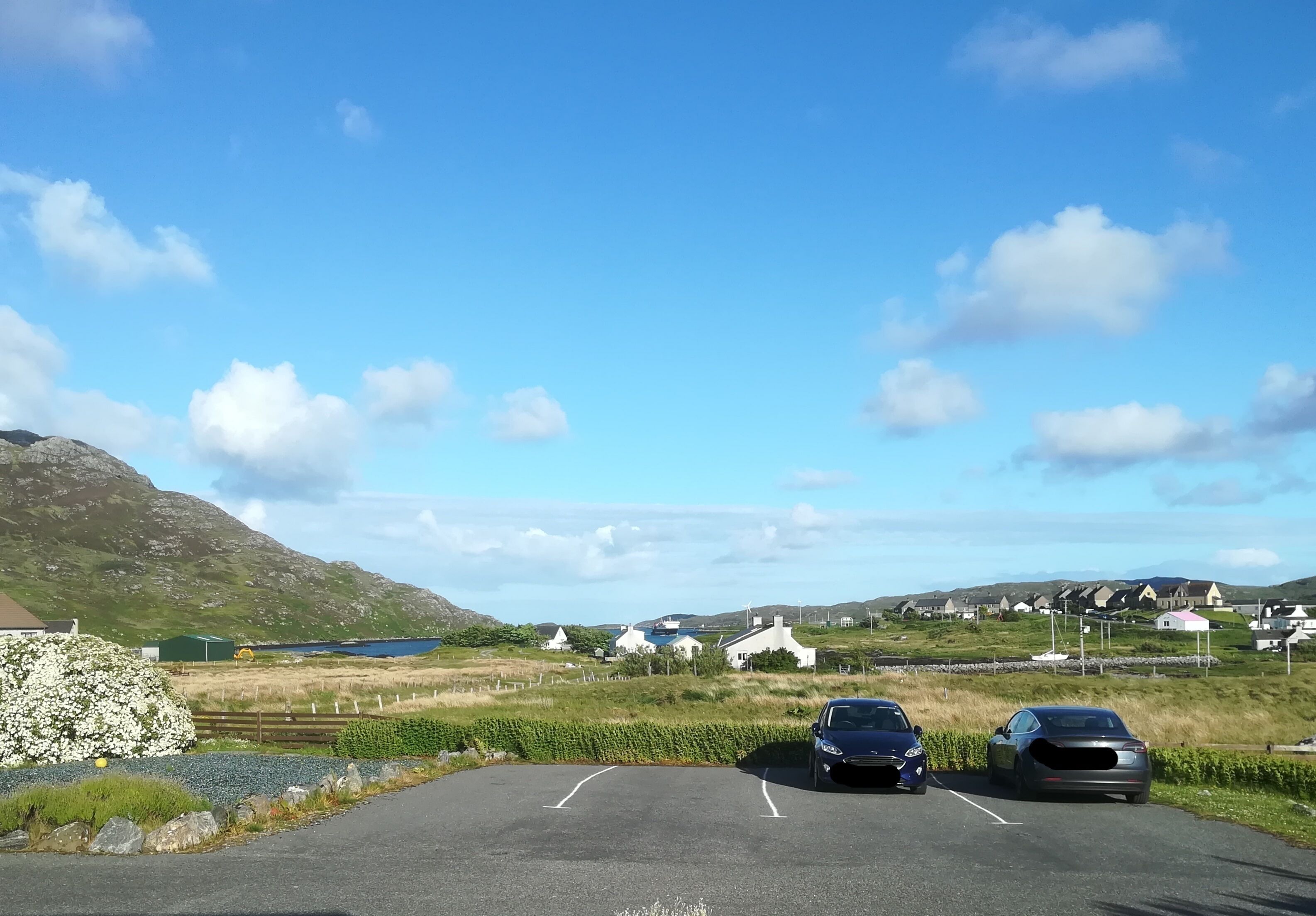 A parking lot with two cars, a stone wall, and houses in the background.