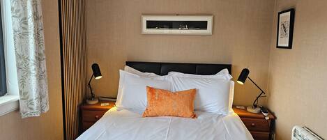 A neatly made bed with white bedding and an orange pillow, flanked by two bedside tables with lamps, in a room with beige walls and a framed picture above the bed.