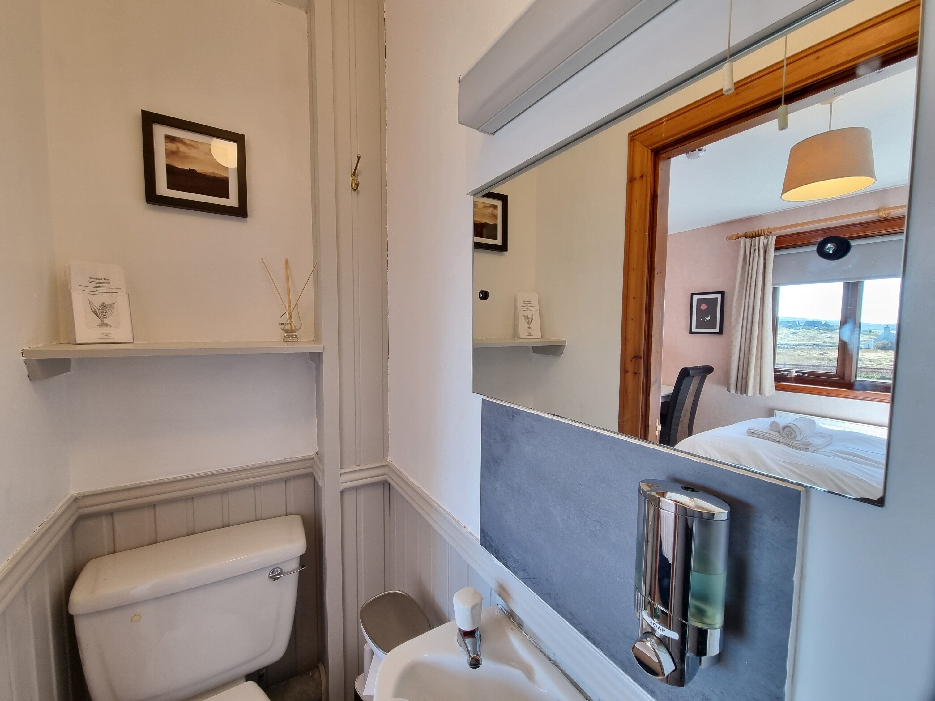 A bathroom with a toilet, sink, and mirror. There are shelves with decorative items and a window with a view.