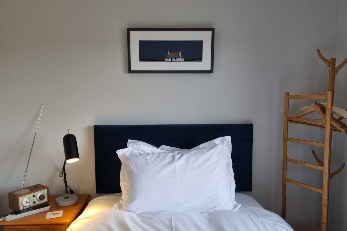 En snyggt bäddad säng med vita sängkläder, en mörk sänggavel, en inramad tavla ovanför, ett träbord vid sängen med en radio och en lampa, och en träklädhängare till höger.