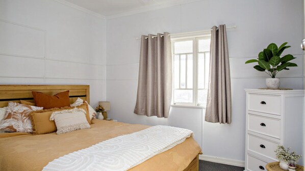 Un dormitorio con cama, mesita de noche, cómoda, ventana con cortinas y una planta en maceta.