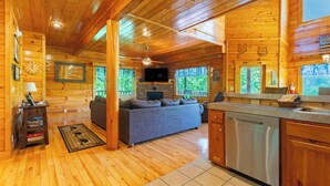 A log cabin interior with a wooden floor, a sofa, a television, a fireplace, and a kitchen area.