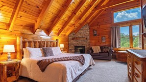 A log cabin bedroom with a large bed, a stone fireplace, a stone wall, a wooden ceiling, and a window with a view of trees.