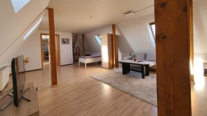 Une chambre spacieuse aménagée en mezzanine, avec une lucarne, des poutres apparentes, un lit, un bureau et un canapé.