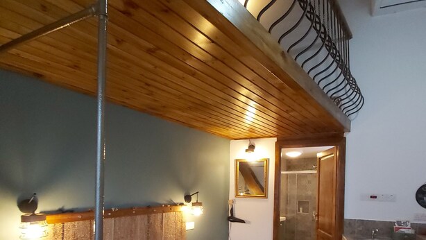 Ein Schlafzimmer mit Holzbalkendecke, einem Bett mit gemusterten Betttextilien, einem Nachttisch, einem Spiegel und einem durchgegangene Tür erkennbaren Badezimmer.