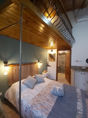Un dormitorio con techo de madera, una cama con ropa de cama estampada, una mesita de noche, un espejo y un baño visible a través de una puerta abierta.