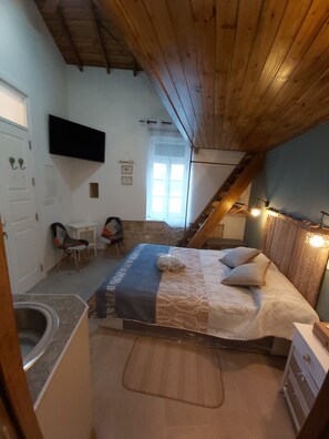 Un dormitorio con cama, escritorio y silla. Se ve una ventana con cortinas, un televisor montado en la pared y techo de madera.