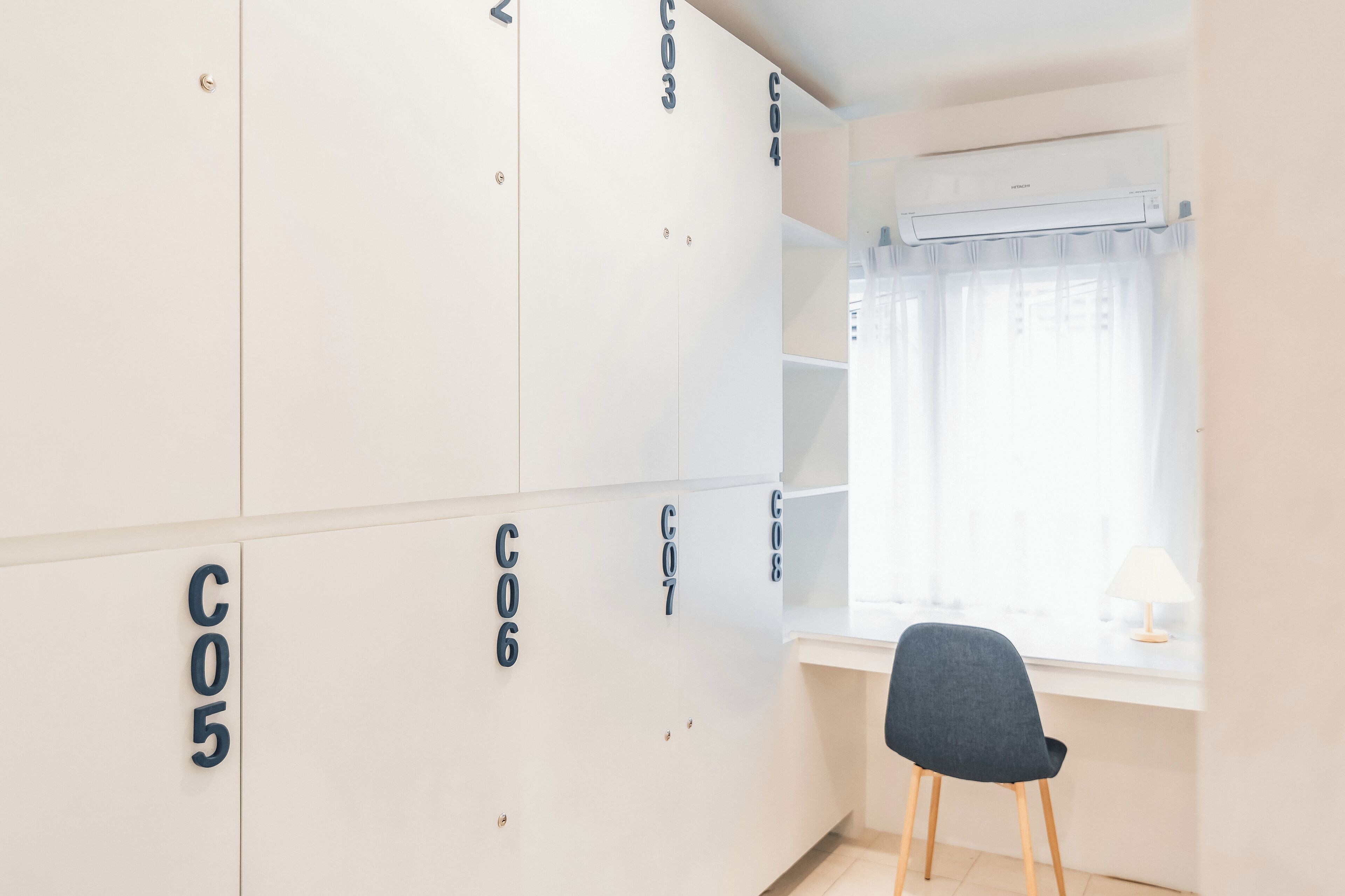 A room with white lockers labeled with numbers, a window with a curtain, and a desk with a lamp.