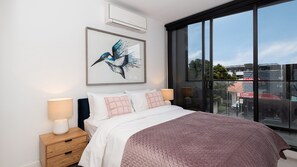 A bedroom with a large bed, a window with a view of buildings, a wall-mounted air conditioner, and a framed artwork of a bird. - Bayside living at The Hampton (Hampton)