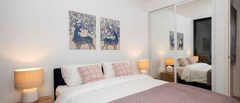 Une chambre à coucher comprenant un lit, deux lampes de chevet, une table de nuit, un grand miroir et deux œuvres d’art mural.