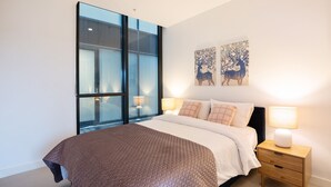 A modern bedroom with a large window, a bed with a brown quilt, two wall-mounted lamps, and two abstract paintings.