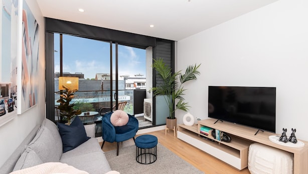 A modern living room with a large TV, comfortable seating, a view of the city through large windows, and a balcony with a plant.