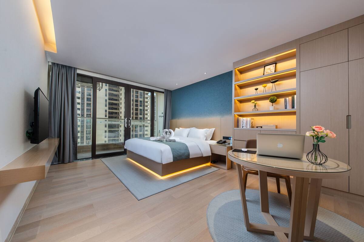 A modern hotel room with a large bed, a desk with a laptop, a TV, and a bookshelf.